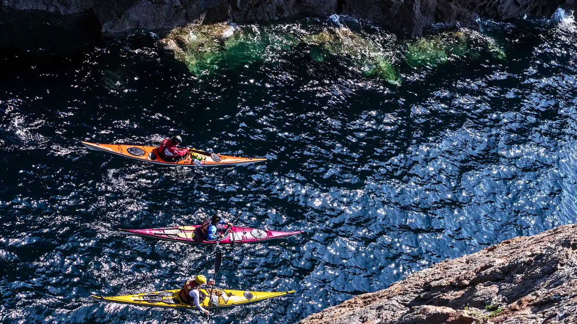 Tres kayakistas de mar navegando en fila cerca de un acantilado rocoso en aguas cristalinas